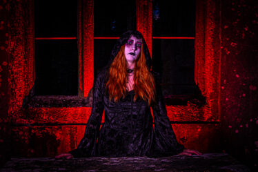 Woman in a black hooded dress with dramatic makeup, posed against a red-lit stone wall and windows at night