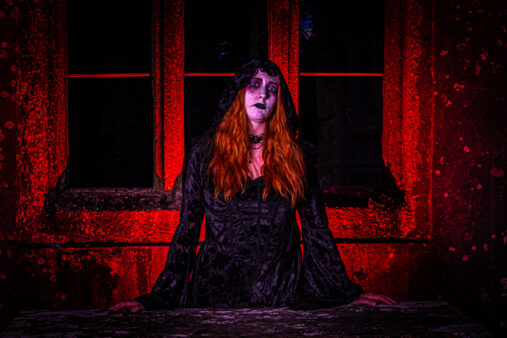 Woman in a black hooded dress with dramatic makeup, posed against a red-lit stone wall and windows at night