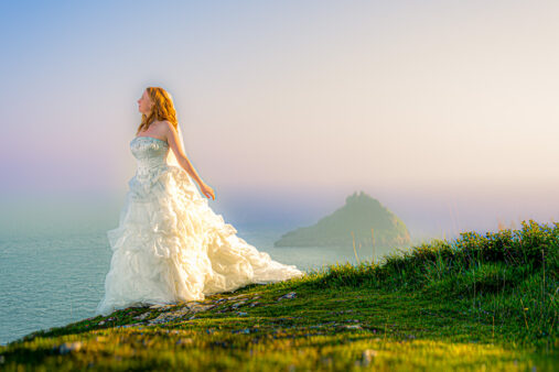 Woman in a white wedding dress standing on a grassy cliff overlooking the sea at sunset, with an island in the distance