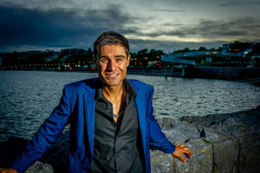 Smiling man in a blue blazer leaning on a stone wall beside the water at dusk, with city lights and a bridge in the background
